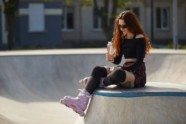 轮滑手女人坐在滑板公园的斜坡顶上 手里拿着玻璃瓶水 年轻酷的滑板手 脚上穿着时髦的内联滑板 — 图库照片