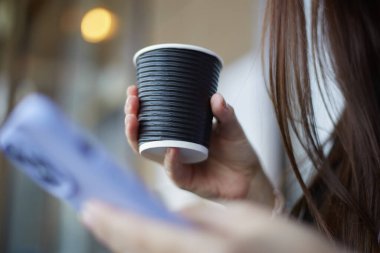 Elinde tek kullanımlık bir fincan kahve tutan genç bir kadın. Bir kadın latte içiyor ve akıllı bir telefonla sosyal medya uygulamasına göz atıyor.