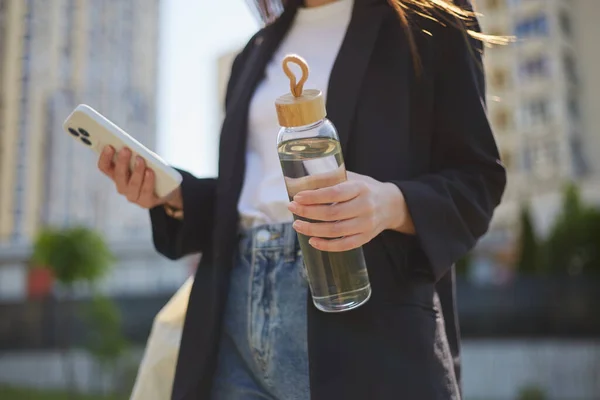 年轻姑娘手里拿着玻璃瓶水和一部现代智能手机 带着小玩意和可重复使用的水瓶在城市街道上行走的难以辨认的女性 — 图库照片
