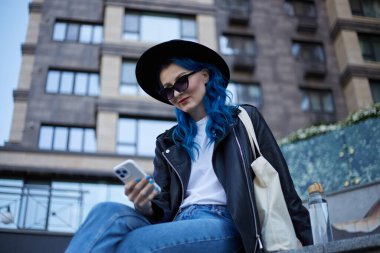 Boyalı mavi saçlı güzel beyaz bir kadın yüksek binadaki bankta oturuyor ve bir cep telefonu uygulamasını inceliyor. Çeşitlilik ve iletişim konsepti