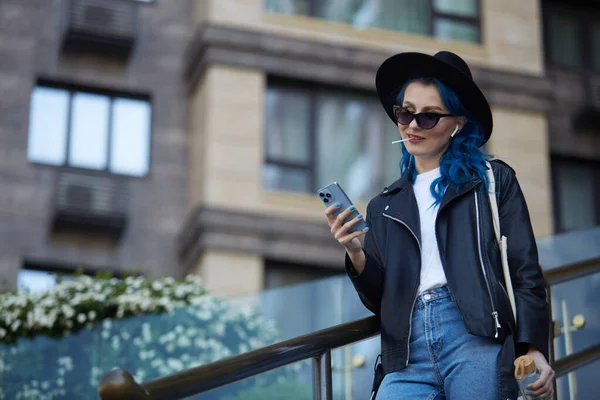 一位漂亮的 多样化的年轻女子 她用智能手机染蓝了头发 一边散步一边用无线耳机听音乐 身着时髦休闲装的白人女性在城市街道上漫步 — 图库照片
