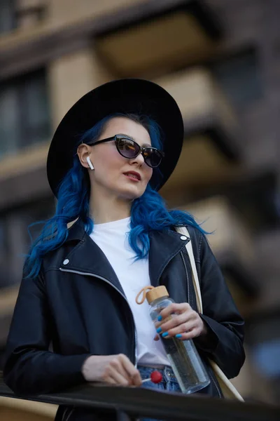 散歩中のワイヤレスヘッドフォンで音楽を聴くサングラスの美しい若い女性の肖像画 街の通りにポーズをとっている青の髪を持つスタイリッシュかつ多様な女性 — ストック写真