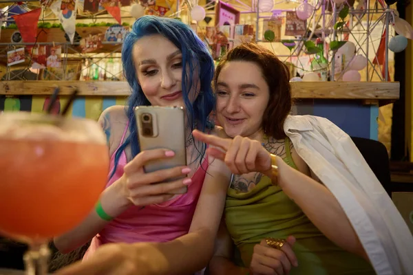 快乐的男女同性恋夫妇在酒吧里玩得开心 两个有着纹身的年轻而多样化的女人带着微笑在酒吧里浏览智能手机应用程序 — 图库照片