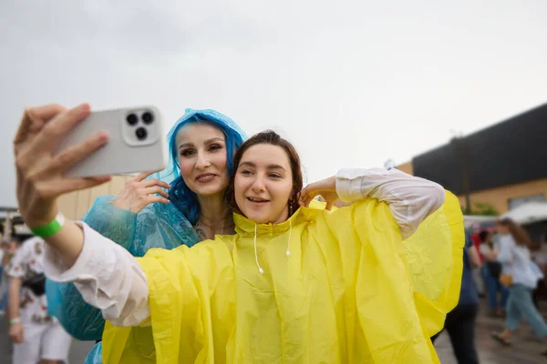 两个穿着雨衣的不同的年轻女子在夏天的节日摆出一张自拍照的姿势 一对快乐的女同性恋 男同性恋 双性恋和变性者夫妇在雨天玩乐的肖像 — 图库照片