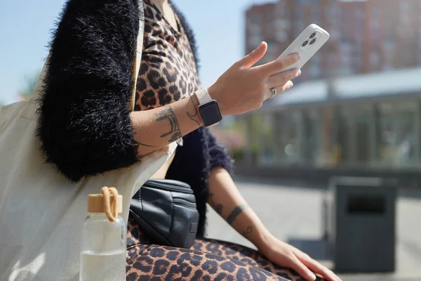 一个时髦的年轻女人坐在长椅上 在一个现代智能手机上用三重相机浏览一个移动应用程序 穿着豹衣 用手机进行交流的无法辨认的女性 — 图库照片