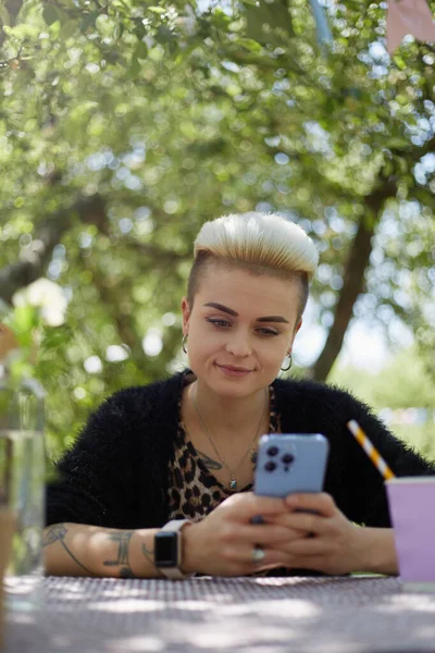 一个时髦的多样化女人 短发坐在一个绿色的公园里 在智能手机上输入一个信息 一个有着汤姆男孩式发型的漂亮白人女性的画像 — 图库照片
