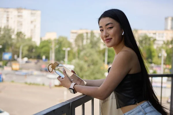 優しい笑顔で屋外でポーズする美しいベトナムの女性の人 ワイヤレスヘッドフォンを装着した陽気なPoc女性の肖像画と手にガラス水ボトルを保持 — ストック写真