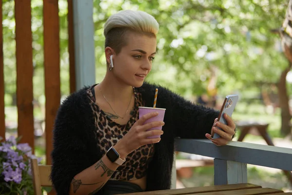 快乐的年轻女人 留着短发 坐在咖啡店里 笑着浏览手机 喝着鸡尾酒 一个有汤姆男孩发型的时髦女性的肖像 — 图库照片