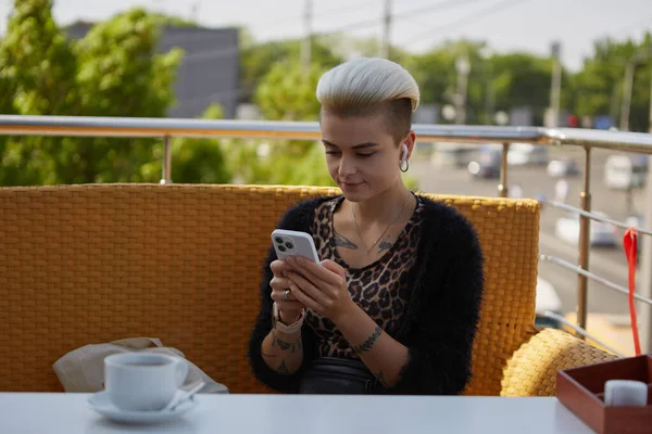 年轻而多样化的女人坐在咖啡店的阳台上 用智能手机输入信息 一个有着短短的漂白头发和纹身的漂亮女性的画像 — 图库照片
