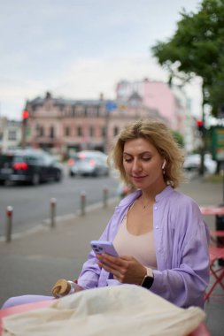 Sokak kafesinde bir masanın arkasında oturan ve akıllı bir telefona mesaj yazan dalgalı saçlı genç sarışın kadın.