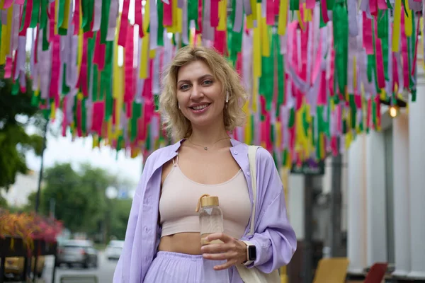 一个快乐的年轻女子站在装饰过的街道中央 手里拿着玻璃瓶水的画像 30多岁时友善的乌克兰女性散步 — 图库照片
