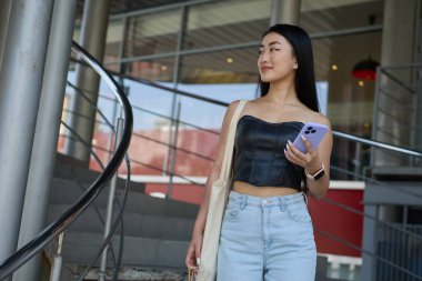 Şık, Asyalı bir kadın elinde akıllı bir telefonla alışveriş merkezinde yürüyor. Neşeli Vietnamlı kadın çantasıyla alışverişe çıktı.