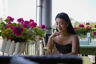 Güzel bir Asyalı kadın açık hava restoranında terasta oturuyor ve bir cep telefonunun menüsünü karıştırıyor. Bir kafede telefon kullanan çekici POC kadın.