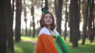 Happy little girl celebrating St Patricks Day. Slow motion video clip of cheerful white kid dressed as fairy and covered with flag of Ireland spinning around in the forest
