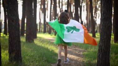 Little girl celebrates St Patrick's Day. Cute 9 year old child in a green pixie costume running through a forest with an Irish flag in slow motion
