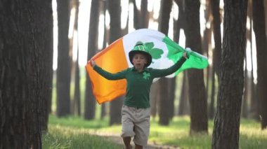 Cute little boy dressed as a leprechaun on St Patricks Day. 10 yeard old kid in green clothes with shamrocks running in the forest with a large Irish flag in hands
