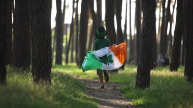 Little boy celebrates Saint Patricks Day. Cute white kid dressed as a leprechaun running with an Irish flag in the forest in slow motion