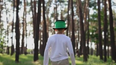 Little boy in green leprechaun hat walking alone through a forest at sunset. White kid dressed for a St Patrick's Day celebration
