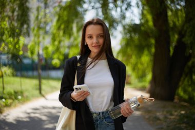 Parkta elinde modern bir akıllı telefon ve cam su şişesiyle yürüyen sevimli genç bir kadın.. 