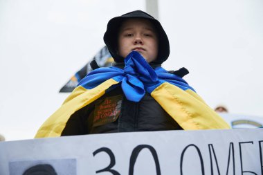 Kayıp askerlere adanmış halka açık bir gösteride omuzlarında ulusal mavi ve sarı bir bayrak taşıyan üzgün Ukraynalı çocuk. Kyiv - 25 Şubat 2024