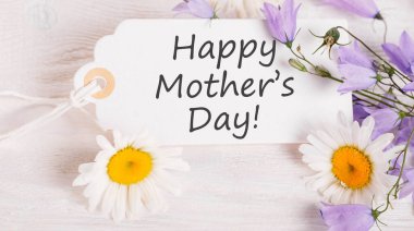 Beyaz papatyalar ve mor çiçeklerle çevrili Anneler Günü için zarif senaryoları olan beyaz not anneler günü mesajı sıcaklığı, beyaz arkaplanı ve saflığı vurguluyor, sevgi dolu bir kutlama için mükemmel.