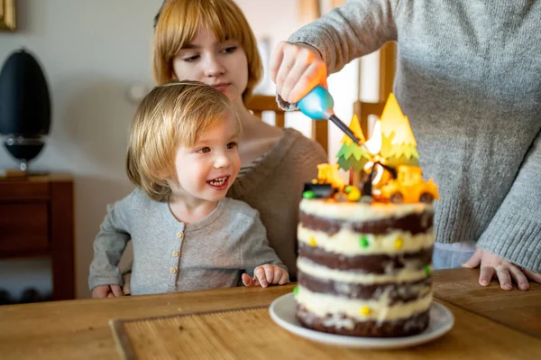 2人の姉が幼児の弟の誕生日ケーキにろうそくを灯している 自家製の誕生日ケーキを切る 自宅で2歳の誕生日を祝う — ストック写真
