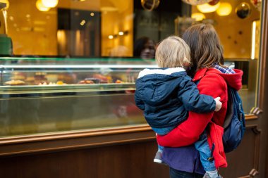 Milan 'ın kuzeydoğusundaki Bergamo şehrinin ortaçağ caddesinde fırın penceresine bakarken kucağında küçük oğlunu tutan genç bir anne. Şehrin üst bölgesi Citta Alta 'nın manzarası. Bergamo, Lombardy, İtalya.