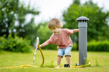 Cute toddler boy watering flower beds in the garden at summer day. Child using garden hose to water vegetables. Kid helping with everyday chores. Mommys little helper.