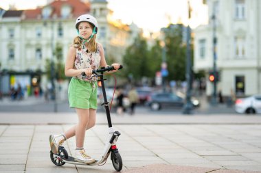 Adorable young girl riding her scooter in a city on sunny summer evening. Pretty preteen child riding a roller. Active leisure and outdoor sports for kids. clipart