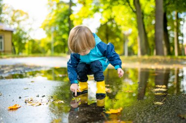 Güneşli bir sonbahar gününde şehir parkında su birikintisinde oynayan sarı lastik çizme giyen sevimli çocuk. Doğayı keşfeden çocuk. Küçük çocuklar için eğlenceli sonbahar aktiviteleri.