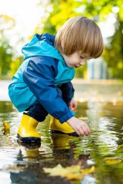 Güneşli bir sonbahar gününde şehir parkında su birikintisinde oynayan sarı lastik çizme giyen sevimli çocuk. Doğayı keşfeden çocuk. Küçük çocuklar için eğlenceli sonbahar aktiviteleri.