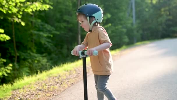 夏の日に赤ちゃんのスクーターに乗っているおかしい幼児の少年 市立公園のミニバイクでのキッズトレーニングバランス 自然を探検する子供 小さい子供のための夏の屋外活動 スローモーション映像 — ストック動画