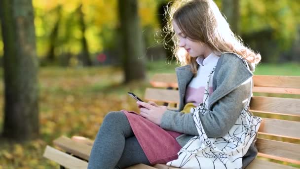 秋天阳光灿烂的一天 漂亮的少女坐在公园的长椅上 检查她的智能手机 在网上冲浪 一个女孩用智能手机拍摄的慢镜头 — 图库视频影像