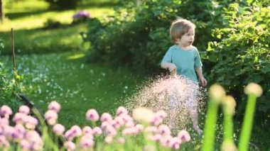 Cute toddler boy watering flower beds in the garden at summer day. Child using garden hose to water vegetables. Kid helping with everyday chores. Mommys little helper.