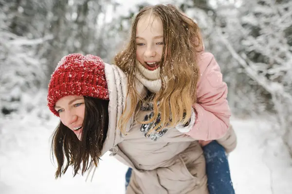 Cute teen girl and her mother having fun on a walk in snow covered pine forest on chilly winter day. Teenage child exploring nature. Winter activities for kids.