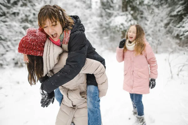 Funny teen sisters and their mother having fun on a walk in snow covered pine forest on chilly winter day. Teenage girls exploring nature. Winter activities for kids.