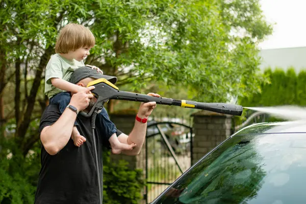 Father and his toddler son using a water gun to wash a car. Male driver washing a car with contactless high pressure water jet. Cleaning a vehicle in a backyard.