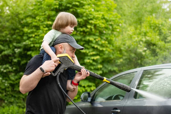 Father and his toddler son using a water gun to wash a car. Male driver washing a car with contactless high pressure water jet. Cleaning a vehicle in a backyard.
