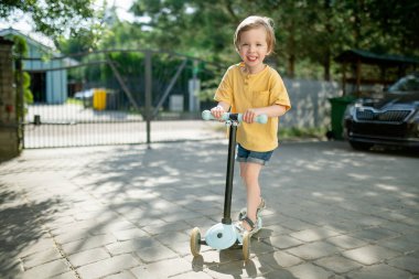 Güneşli bir yaz akşamında arka bahçede scooterını süren sevimli küçük bir çocuk. Paten süren küçük bir çocuk. Çocuklar için aktif eğlence ve açık hava sporları.