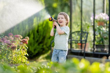 Yaz günü bahçede çiçek tarhlarını sulayan tatlı bir çocuk. Çocuk sebzeleri sulamak için bahçe hortumu kullanıyor. Çocuk günlük ev işlerine yardım ediyor. Annesinin küçük yardımcısı..
