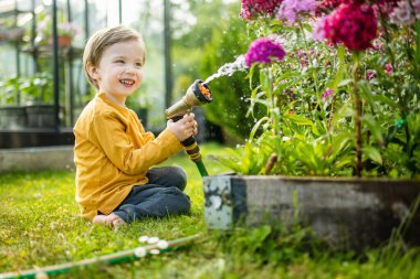 Yaz günü bahçede çiçek tarhlarını sulayan tatlı bir çocuk. Çocuk sebzeleri sulamak için bahçe hortumu kullanıyor. Çocuk günlük ev işlerine yardım ediyor. Annesinin küçük yardımcısı..