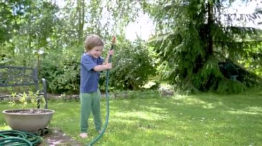 Yaz günü bahçede çiçek tarhlarını sulayan tatlı çocuk. Çocuk sebzeleri sulamak için bahçe hortumu kullanıyor. Çocuk günlük ev işlerine yardım ediyor. Sıcak güneşli yaz günü açık hava eğlencesi. Yavaş çekim.