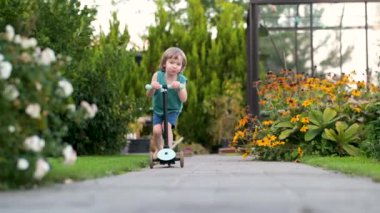 Güneşli bir yaz gününde arka bahçede scooterını süren sevimli küçük bir çocuk. Paten süren küçük bir çocuk. Çocuklar için aktif eğlence ve açık hava sporları. Yavaş çekim görüntüleri.