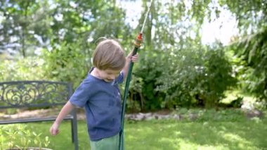 Yaz günü bahçede çiçek tarhlarını sulayan tatlı çocuk. Çocuk sebzeleri sulamak için bahçe hortumu kullanıyor. Çocuk günlük ev işlerine yardım ediyor. Sıcak güneşli yaz günü açık hava eğlencesi. Yavaş çekim.