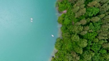 Tropikal göl kıyısı boyunca giden teknelerin yukarıdan aşağıya doğru dönen görüntüsü. Çam ormanlarıyla çevrili manzaralı zümrüt göl.