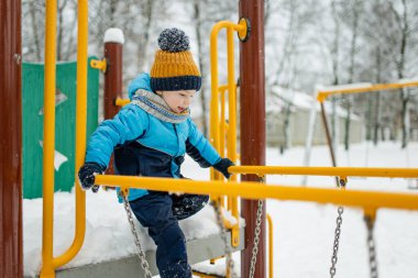 Kar yağışlı kış gününde oyun parkında eğlenen sevimli küçük çocuk. Karda oynayan, kalın giysiler giymiş şirin bir çocuk. Çocuklu aileler için kış etkinlikleri.
