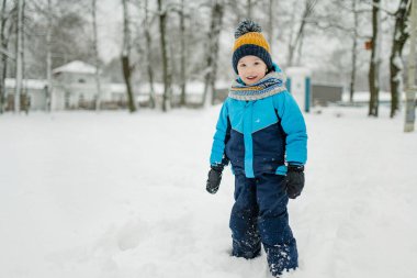 Sevimli küçük çocuk karlı kış gününde eğleniyor. Karda oynayan, kalın giysiler giymiş şirin bir çocuk. Çocuklu aileler için kış etkinlikleri.