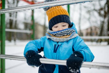 Kar yağışlı kış gününde oyun parkında eğlenen sevimli küçük çocuk. Karda oynayan, kalın giysiler giymiş şirin bir çocuk. Çocuklu aileler için kış etkinlikleri.