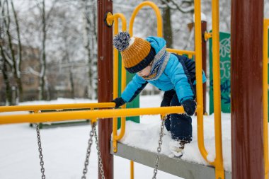 Kar yağışlı kış gününde oyun parkında eğlenen sevimli küçük çocuk. Karda oynayan, kalın giysiler giymiş şirin bir çocuk. Çocuklu aileler için kış etkinlikleri.