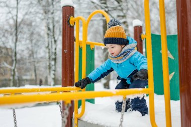 Kar yağışlı kış gününde oyun parkında eğlenen sevimli küçük çocuk. Karda oynayan, kalın giysiler giymiş şirin bir çocuk. Çocuklu aileler için kış etkinlikleri.